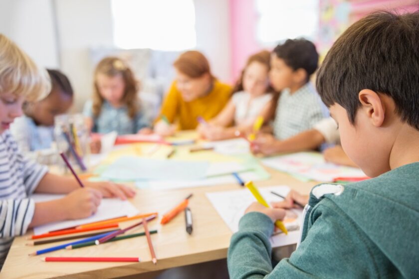 Children sitting at a table, focused on drawing with colored pencils and markers on paper. An adult is assisting in the background.